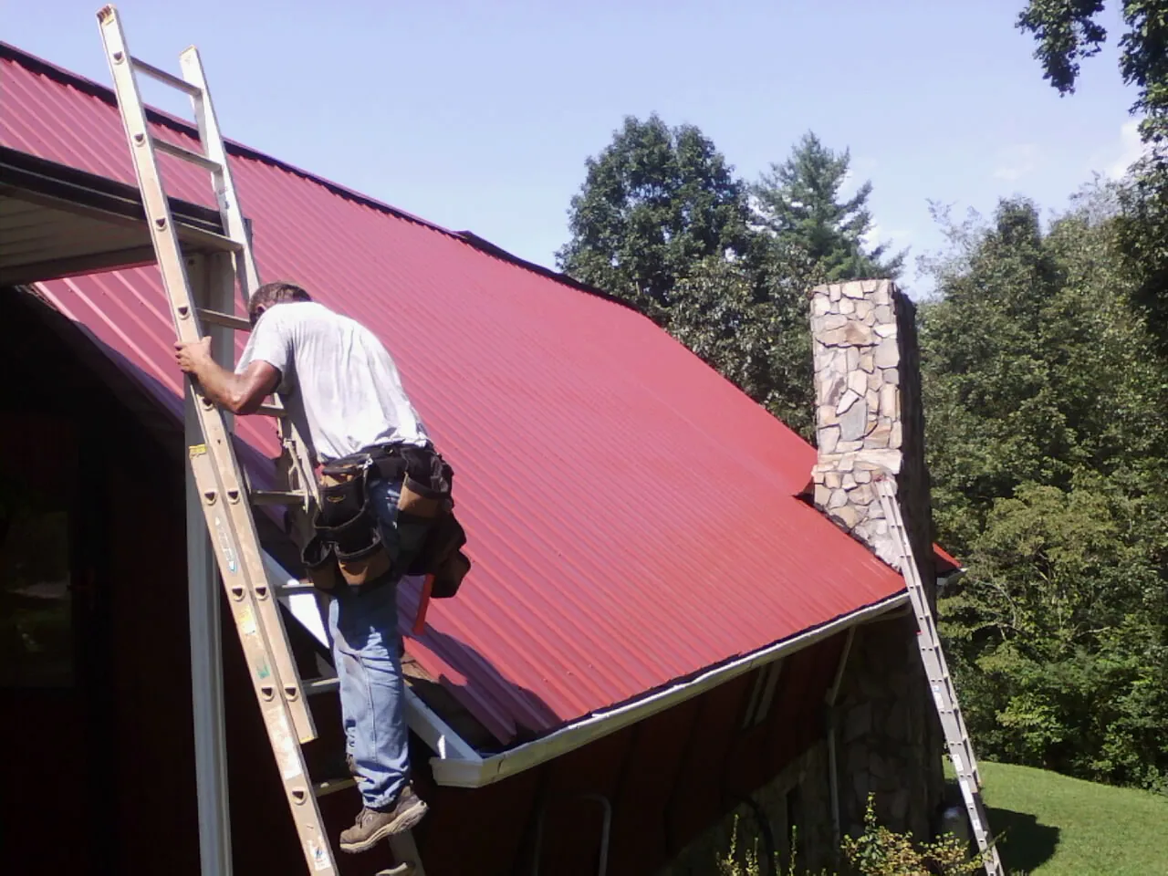 Roofer on a ladder beside a red metal roof and stone chimney — Soffit & Fascia Repair by Roofing Brothers in Westernport